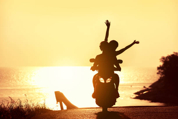 unrecognizable couple with raised arms rides on scooter against sunset sea
